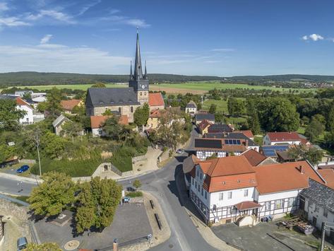 Drohnenaufnahme des Dorfplatzes von Neunhofen mit einer Kirche im Hintergrund und Altbauten im Vordergrund. Das Bild ist im Sommer um die Mittagszeit bei strahlendem Sonnenschein entstanden.