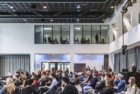 Seitenaufnahme des großen Veranstaltungssaals. Auf der Hauptbühne findet ein Vortrag statt. Parallel dazu sieht man im ersten Stock durch eine Glasfassade die Teilnehmenden eines Workshops.