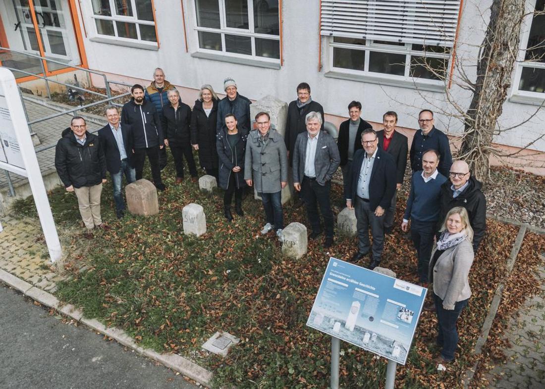 Gruppenfoto aus etwa drei Metern Höhe mit der Drohne aufgenommen. Die Teilnehmer der Veranstaltung stehen nebeneinander auf einer Rasenfläche. Die Grenzsteine des lapidariums der Hohenwindenstraße 14 sind gut dazwischen zu sehen.