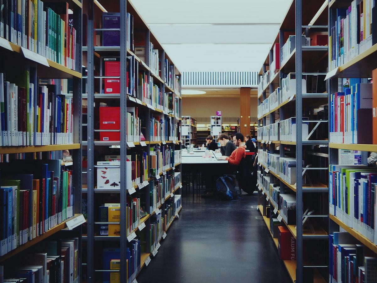 Blickführung durch eine Reihe Bücherregale hin zu Studierenden, die an einem Tisch lesen