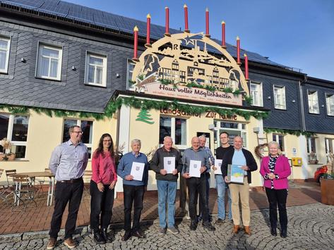 Ein Gruppenfoto der Veranstaltungsteilnehmer:innen vor einem Haus bei Sonnenschein. Der Vorstand hält Urkunden in den Händen.
