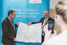 TTG-Geschätfsführer und TLBG-Präsident präsentieren gemeinsam die neue Karte vor der TLBG-Pressewand. Herr Köhler streckt seinen Arm aus in Richtung Karte. Im Vordergrund steht eine junge Frau der TTG und macht ein Handyfoto von der Situation.