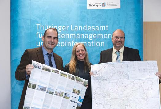 Die drei Hauptakteure des Projekt zeigen jeweils Kartenvorderseite und Kartenrückseite vor der Pressewand des TLBG.