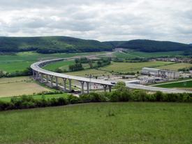Blick von einer Anhöhe in ein Tal, dass von einer geschwungenen Autobahnbrücke durchschnitten wird. Links davon befinden sich Agrarflächen, rechts ist ein Industriegebiet zu sehen. In der Ferne erkennt man eine Ortschaft und weitere Hügel.
