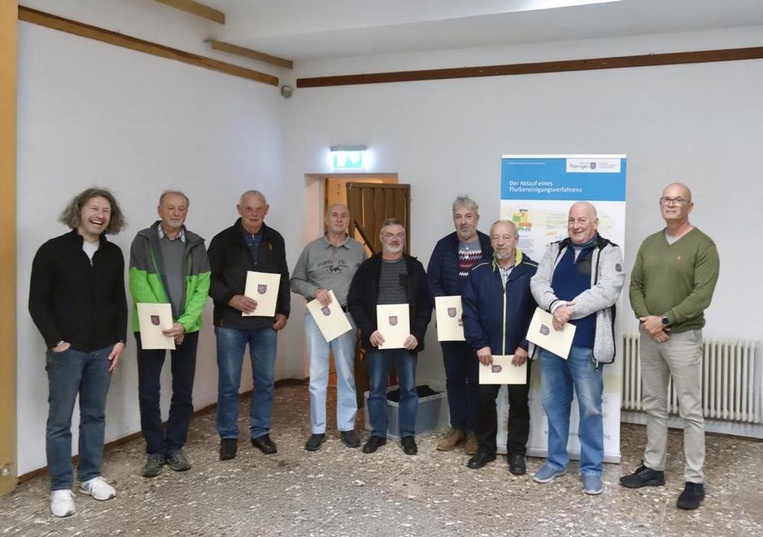 Sieben Männer stehen mit Urkunden in der Hand in einem Raum und blicken zum Fotografierenden. Links und rechts flankieren Kollegen des TLBG. Im Hintergrund aufgebaut ist ein Roll-up mit dem Titel "Ablauf eines Flurbereinigungsverfahrens".