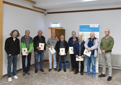 Sieben Männer stehen mit Urkunden in der Hand in einem Raum und blicken zum Fotografierenden. Links und rechts flankieren Kollegen des TLBG. Im Hintergrund aufgebaut ist ein Roll-up mit dem Titel "Ablauf eines Flurbereinigungsverfahrens".