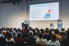 Blick über dutzende Besucher hinweg auf die Hauptbühne mit zwei Referierenden. Auf der Beamerleinwand ist eine Seite der Powerpoint zum Thema KI im Behördenalltag zu sehen.